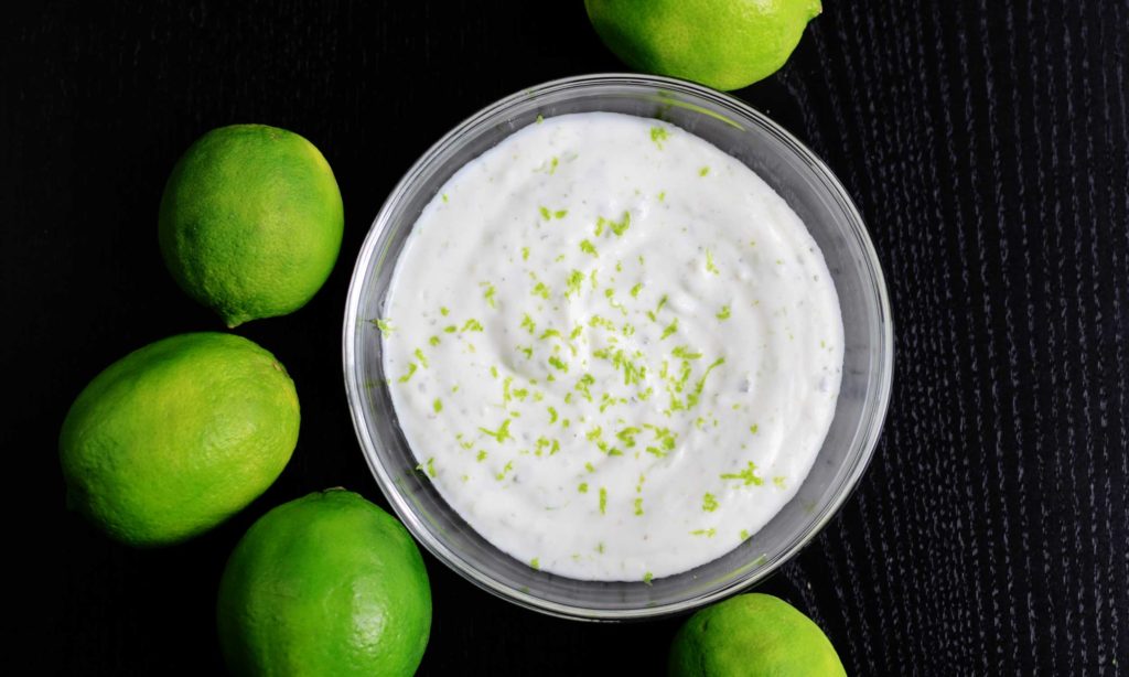 a bowl of lime crema topped with vibrant green lime zest surrounded by fresh limes for this mexican lime crema recipe